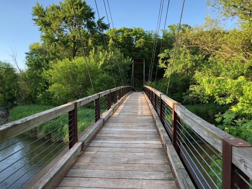 A wooden suspension bridge stretches over a calm waterway, surrounded by lush green trees and foliage.