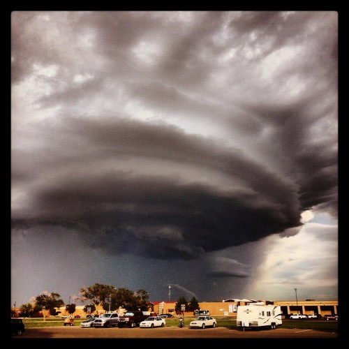 Dramatic storm clouds swirl ominously over a parking lot, casting a dark shadow on nearby buildings and vehicles.