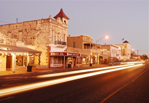 A charming street lined with historic stone buildings, adorned with festive lights, at dusk with light trails from passing cars.