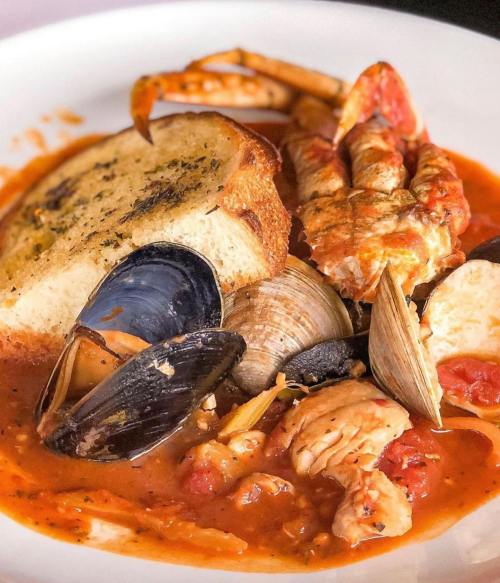 A plate of seafood stew with mussels, clams, lobster, and a slice of garlic bread in a rich tomato broth.