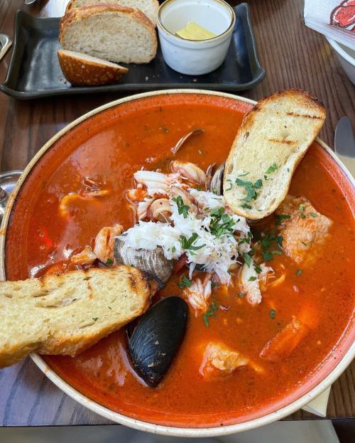 A bowl of vibrant seafood soup with shrimp, clams, and fish, garnished with herbs and served with toasted bread.