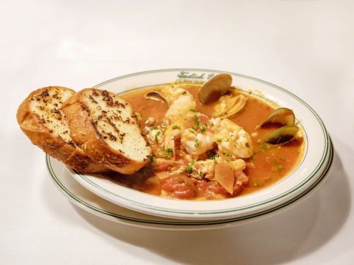 A bowl of seafood stew with shrimp, clams, and tomatoes, served with two slices of garlic bread on the side.