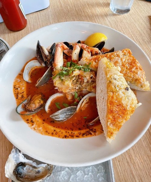 A seafood dish featuring shrimp, mussels, and clams in a rich sauce, served with bread and a lemon wedge.