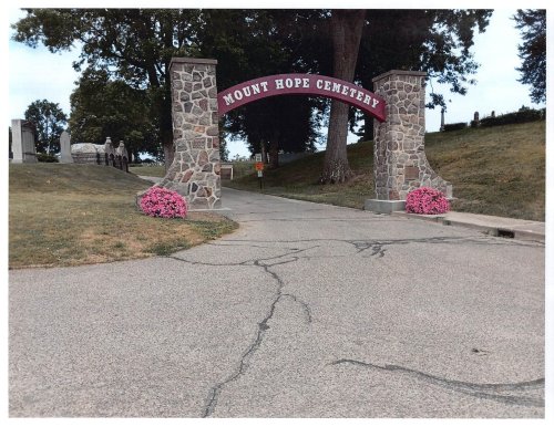 Archway entrance to Mount Hope Cemetery, flanked by pink flowers, with a tree-lined path leading into the cemetery.