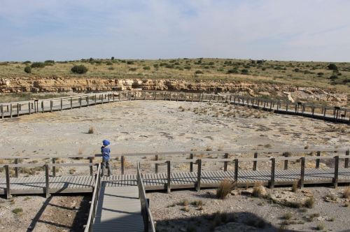 A person stands on a wooden walkway overlooking a dry, rocky landscape with sparse vegetation and distant hills.