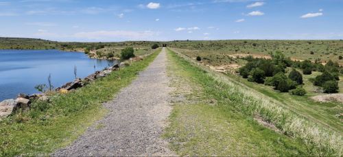 A gravel path runs alongside a calm lake, surrounded by green hills and blue skies with scattered clouds.