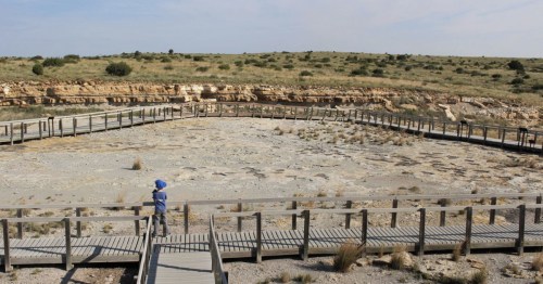 A person stands on a wooden walkway overlooking a dry, rocky landscape with sparse vegetation and distant hills.