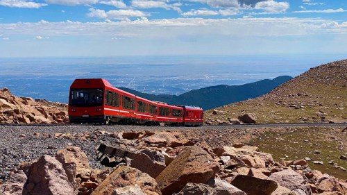 A red train winds along a rocky mountain path with a scenic view of valleys and distant mountains under a blue sky.
