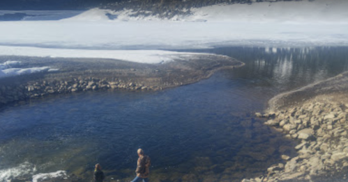 Two people stand by a river with icy banks, surrounded by snow-covered terrain and trees in the background.