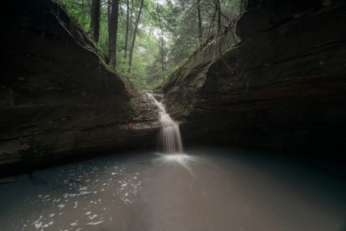 A serene waterfall cascades into a tranquil pool, surrounded by lush green trees and rocky cliffs.