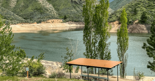 A serene lake surrounded by green hills, with a shaded picnic area in the foreground.