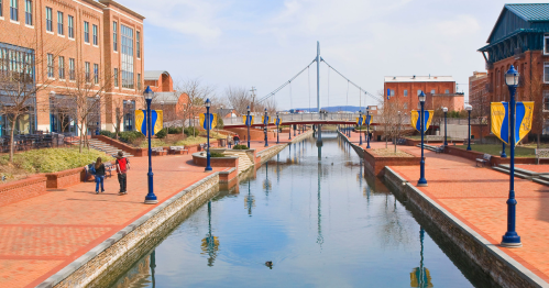 A scenic canal with brick pathways, lined with banners, and buildings in the background under a clear blue sky.