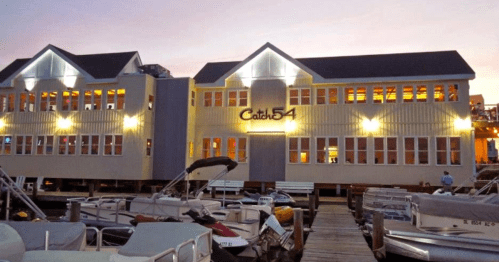 A waterfront restaurant, Catch 54, with bright lights and boats docked in front during twilight.