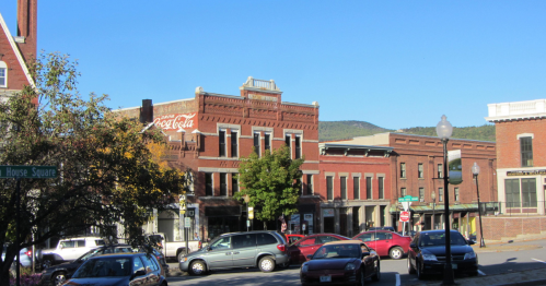 Historic buildings line a street in a small town, with a Coca-Cola mural and parked cars under a clear blue sky.