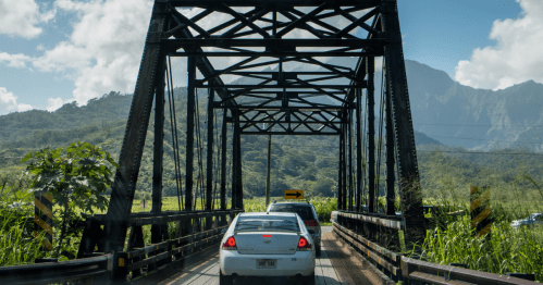 A car drives over a black metal bridge surrounded by lush green mountains and a clear blue sky.