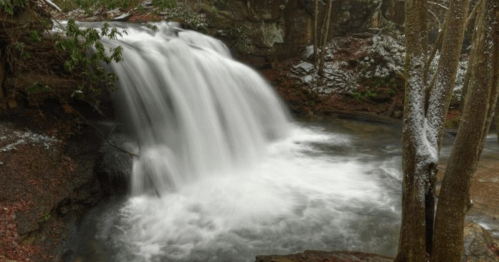 A serene waterfall cascading over rocks, surrounded by trees and a hint of snow on the ground.