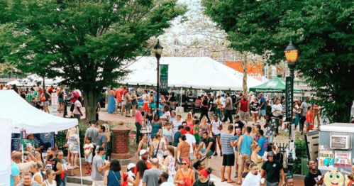 A bustling outdoor market scene with large crowds, tents, and trees, filled with people enjoying the event.