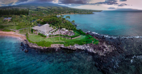 Aerial view of a coastal restaurant on a lush green cliff, surrounded by turquoise waters and rocky shoreline.