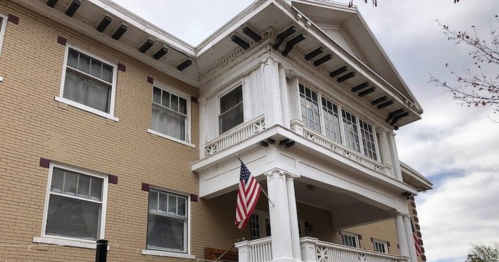 Historic building with a large porch, white trim, and American flags, set against a cloudy sky.
