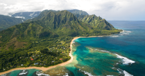 Aerial view of lush green mountains meeting a sandy beach and turquoise ocean under a cloudy sky.