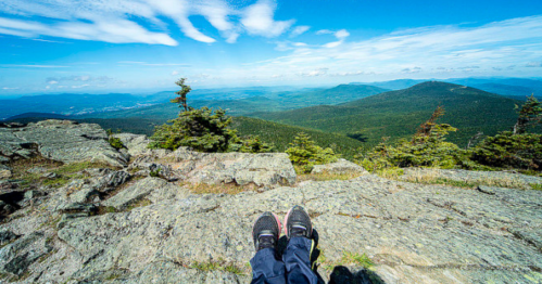 Person's feet resting on a rocky ledge, overlooking a vast green landscape and blue sky with scattered clouds.