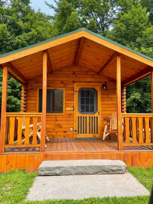 A cozy wooden cabin with a porch, surrounded by lush green trees and a stone pathway leading to the entrance.