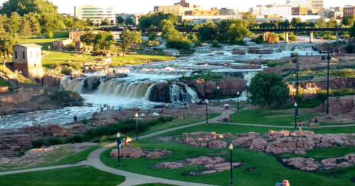 A scenic view of a waterfall cascading over rocks, surrounded by greenery and a park with people enjoying the area.