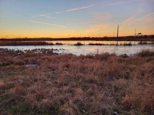 A serene landscape at sunset, featuring a calm water body surrounded by tall grass and distant hills.