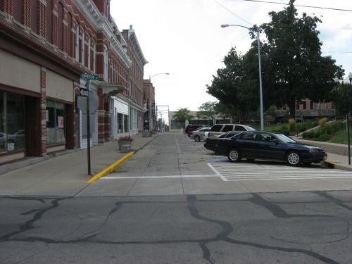 A quiet street scene featuring parked cars, brick buildings, and trees under a partly cloudy sky.