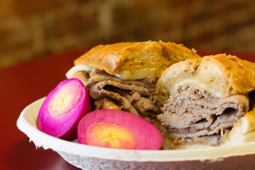 A close-up of a sandwich filled with sliced beef, served with vibrant pink pickled onions on the side.