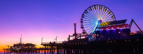 Colorful ferris wheel and pier illuminated at sunset, with vibrant purple and orange sky in the background.