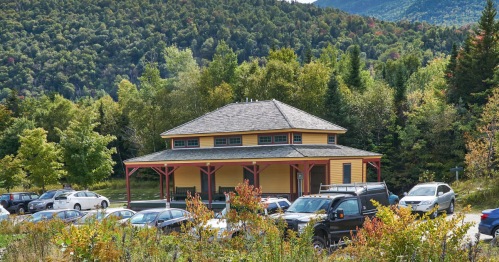 A yellow building with a gray roof surrounded by trees and parked cars, set against a mountainous backdrop.