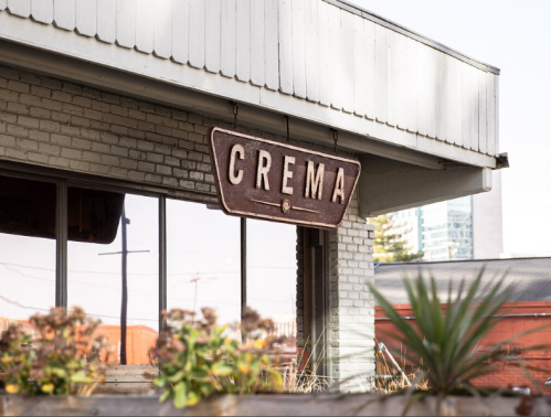 A weathered sign reading "CREMA" hangs from a building, with plants in the foreground and a cityscape in the background.