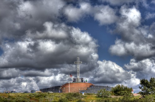 A large cross atop a hill, surrounded by clouds, with flags and visitors in the background.