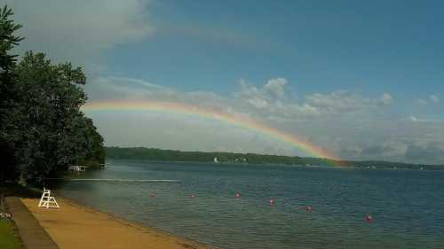 A serene lake scene with a rainbow arching over the water, surrounded by trees and a sandy beach.