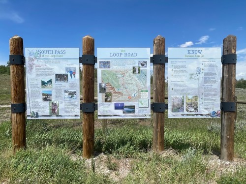 Three informational signs on wooden posts about South Pass, the Loop Road, and visitor guidelines in a grassy area.