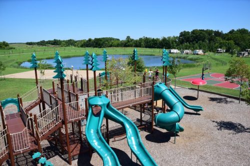 A playground with teal slides, surrounded by green trees and a pond, with basketball courts in the background.