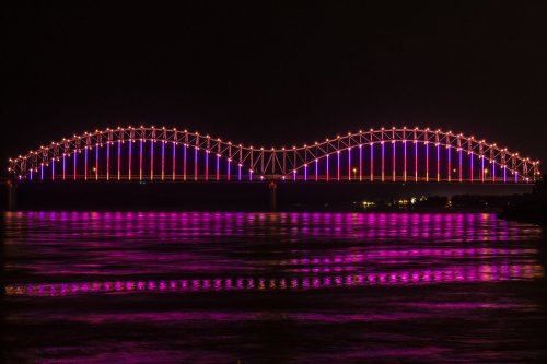 A brightly lit bridge at night, featuring colorful purple and pink lights reflecting on the water below.