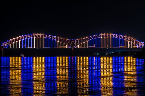 A brightly lit bridge arches over a river at night, reflecting colorful lights on the water's surface.