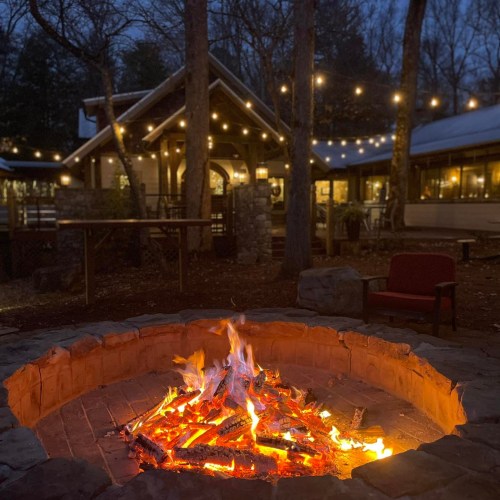 A cozy outdoor fire pit with flames, surrounded by trees and a warmly lit cabin in the background at dusk.