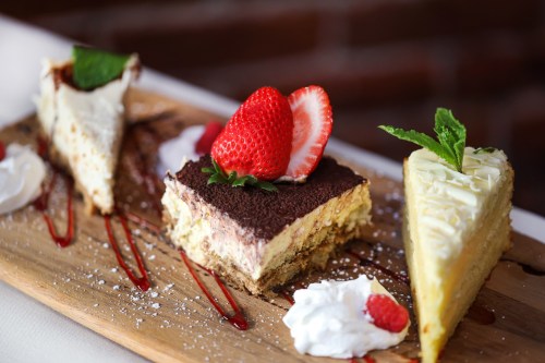 A wooden platter with three dessert slices: tiramisu, cheesecake, and a cream pie, garnished with strawberries and mint.