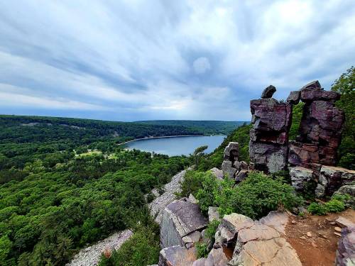 A scenic view from a rocky overlook, showcasing a lush green landscape and a calm lake under a cloudy sky.