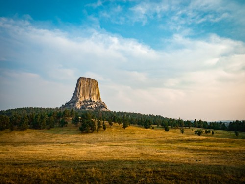 A towering rock formation rises above a grassy landscape, surrounded by trees under a blue sky with soft clouds.