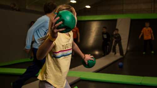 A child in a yellow shirt prepares to throw a green ball in a trampoline park, with other kids playing in the background.