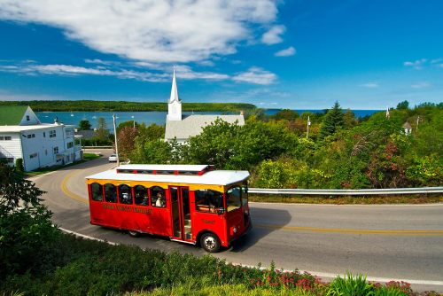 A red trolley bus drives along a scenic road with lush greenery and a church in the background by the water.