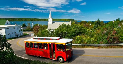 A red trolley bus drives along a scenic road with trees, a church, and a lake in the background under a blue sky.