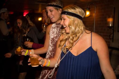 Two women in vintage attire dance joyfully at a lively party, holding drinks and wearing headbands and pearls.
