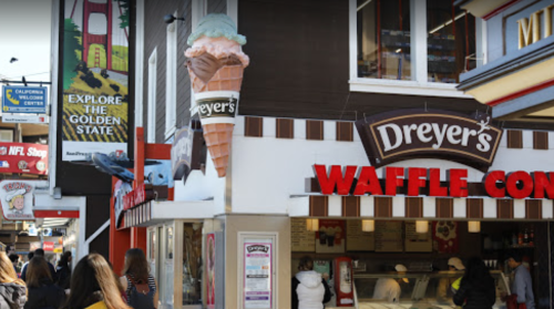 A busy ice cream shop with a large waffle cone sign and people walking by in a vibrant street scene.
