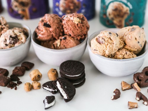 Three bowls of ice cream in various flavors, surrounded by cookies and chocolate pieces on a white surface.