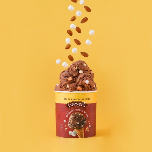 A cup of Dreyer's Rocky Road ice cream topped with almonds and marshmallows, set against a bright yellow background.